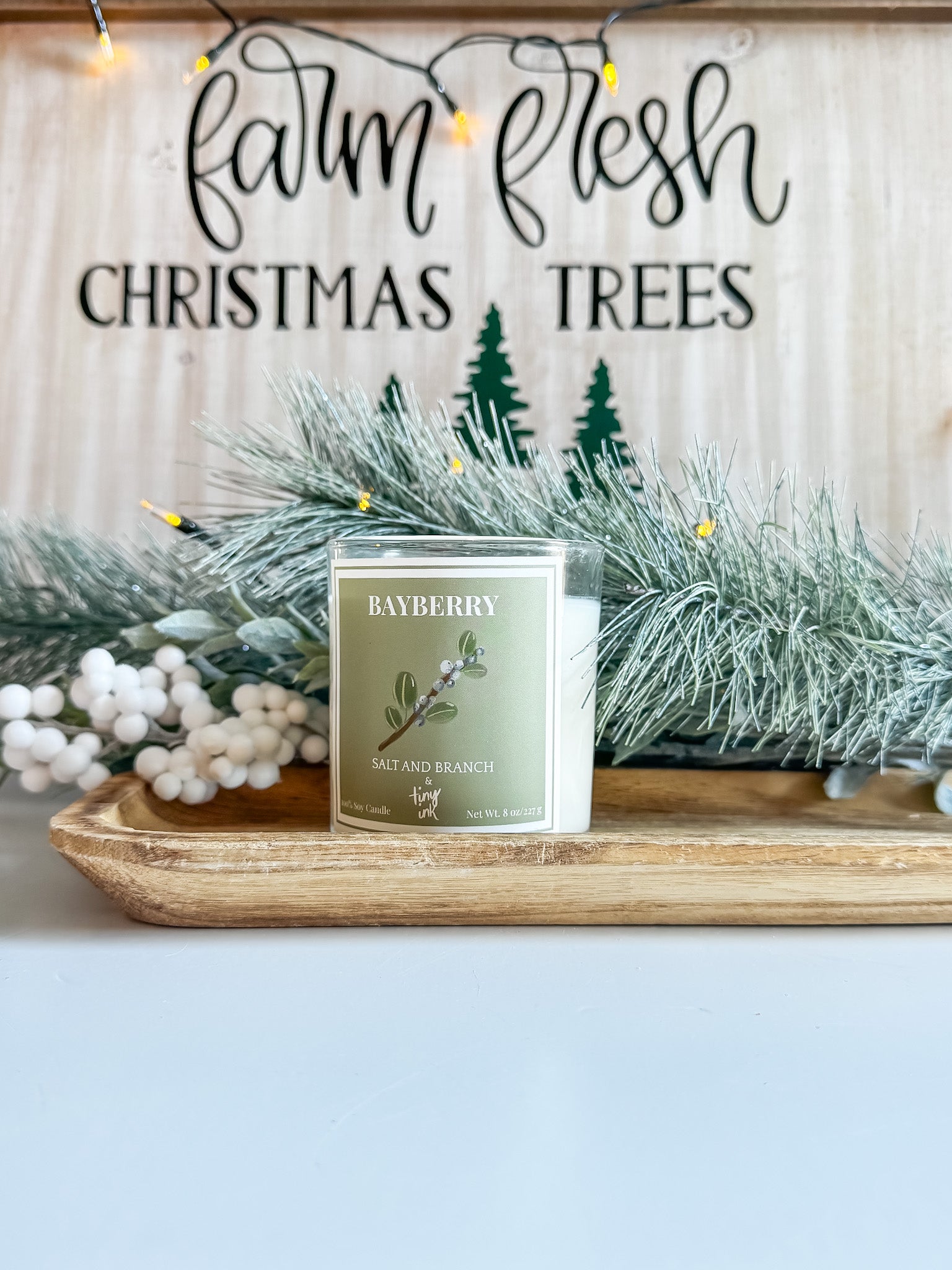 Bayberry candle on a wooden tray with Christmas decorations and 'Farm Fresh Christmas Trees' sign in the background.