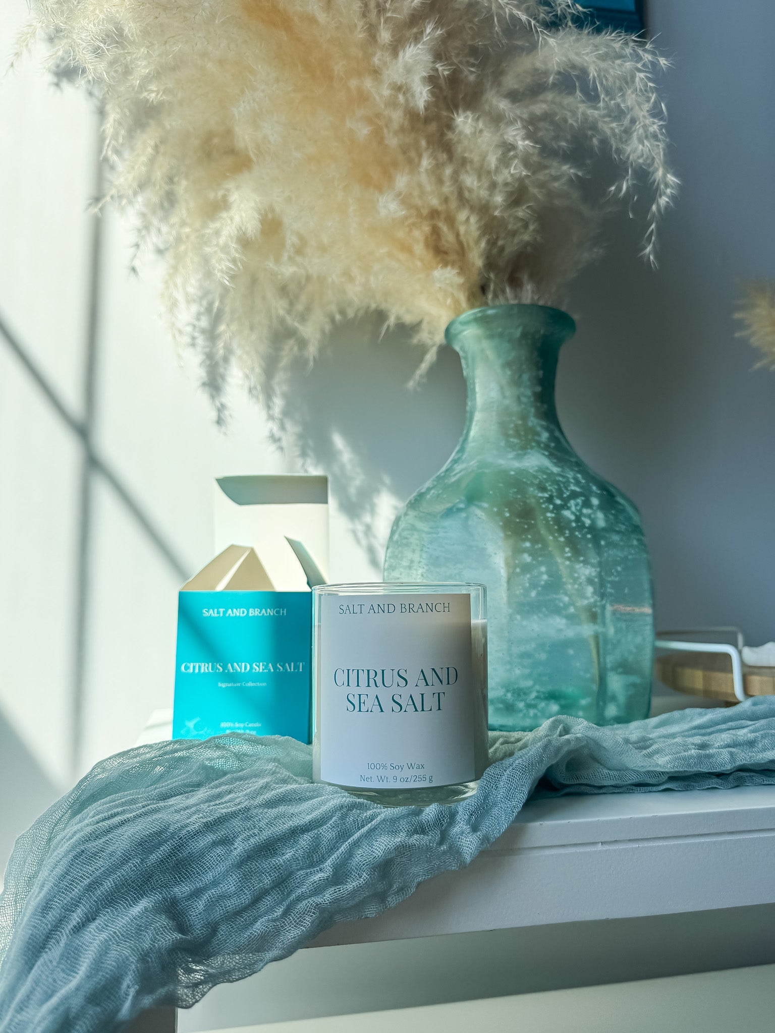 Candle labeled 'Citrus and Sea Salt' with a decorative vase and dried plants on a windowsill. Salt and Branch