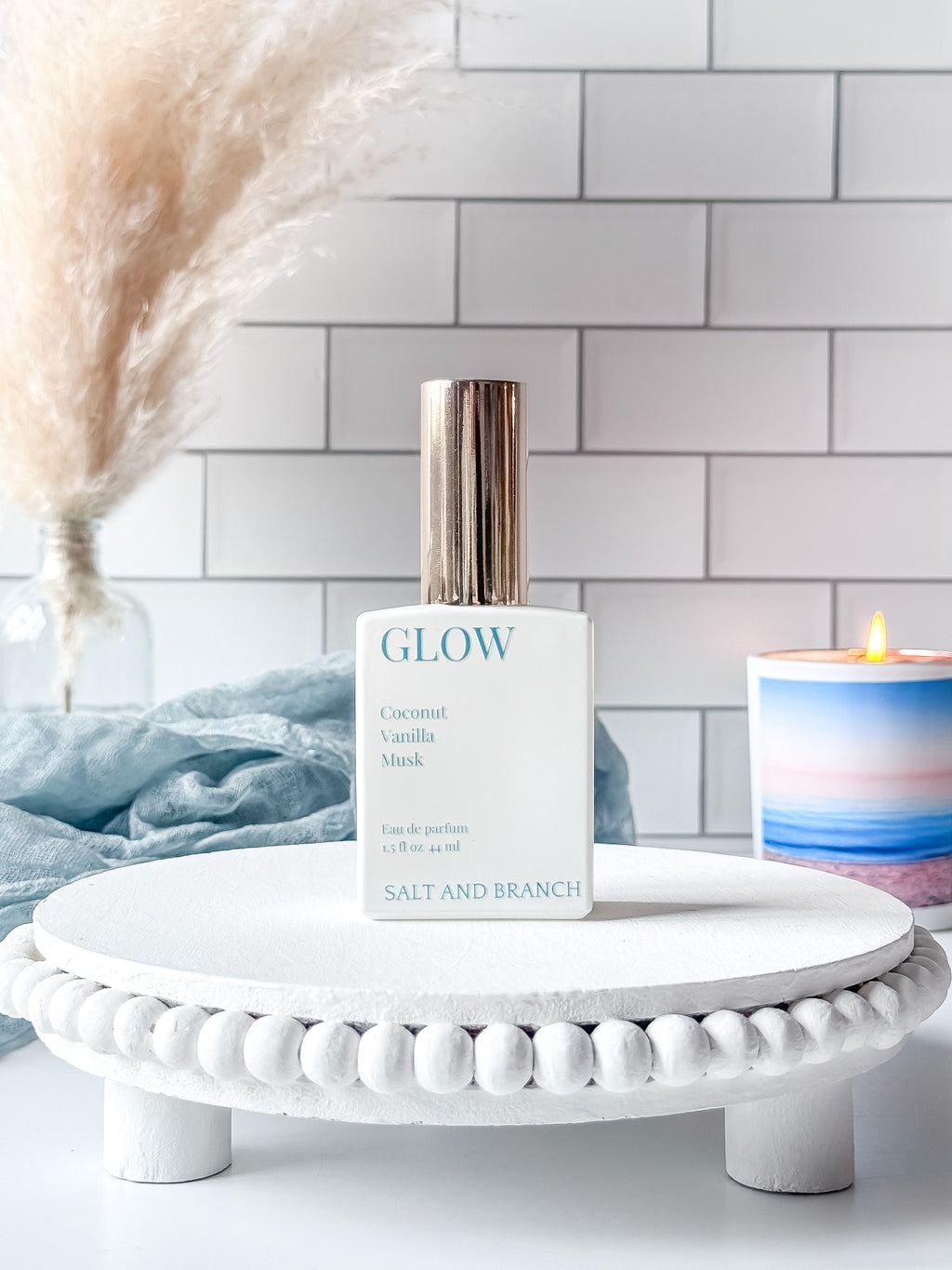 GLOW perfume bottle on a white stand with a candle and decorative elements in the background.