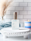 GLOW perfume bottle on a white stand with a candle and decorative elements in the background.