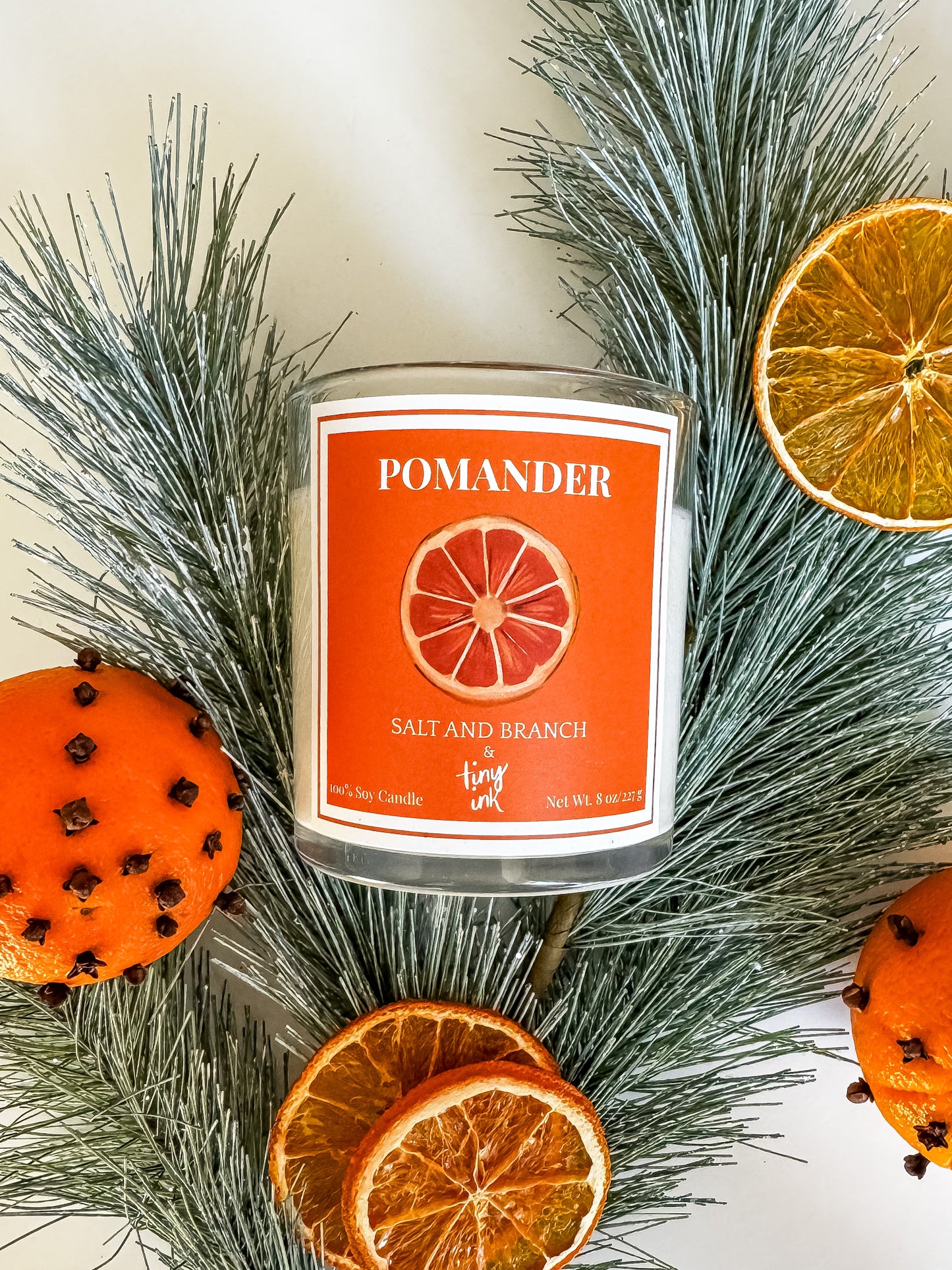 Pomander candle with an orange label surrounded by pine branches and dried oranges.