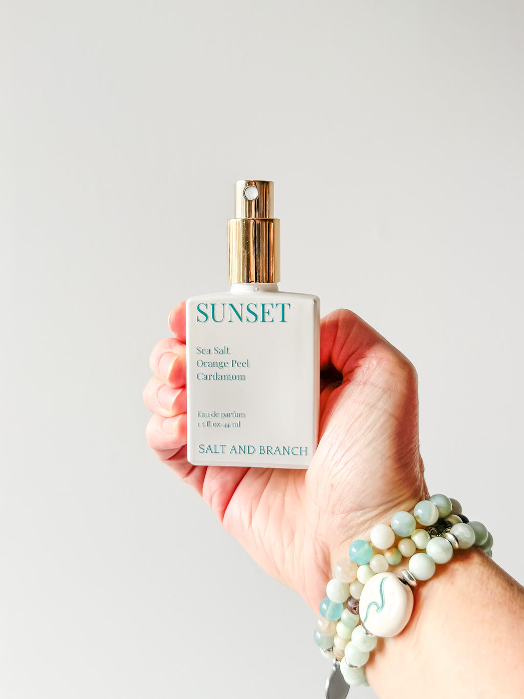 Hand holding a 'Sunset' perfume bottle with a plain background