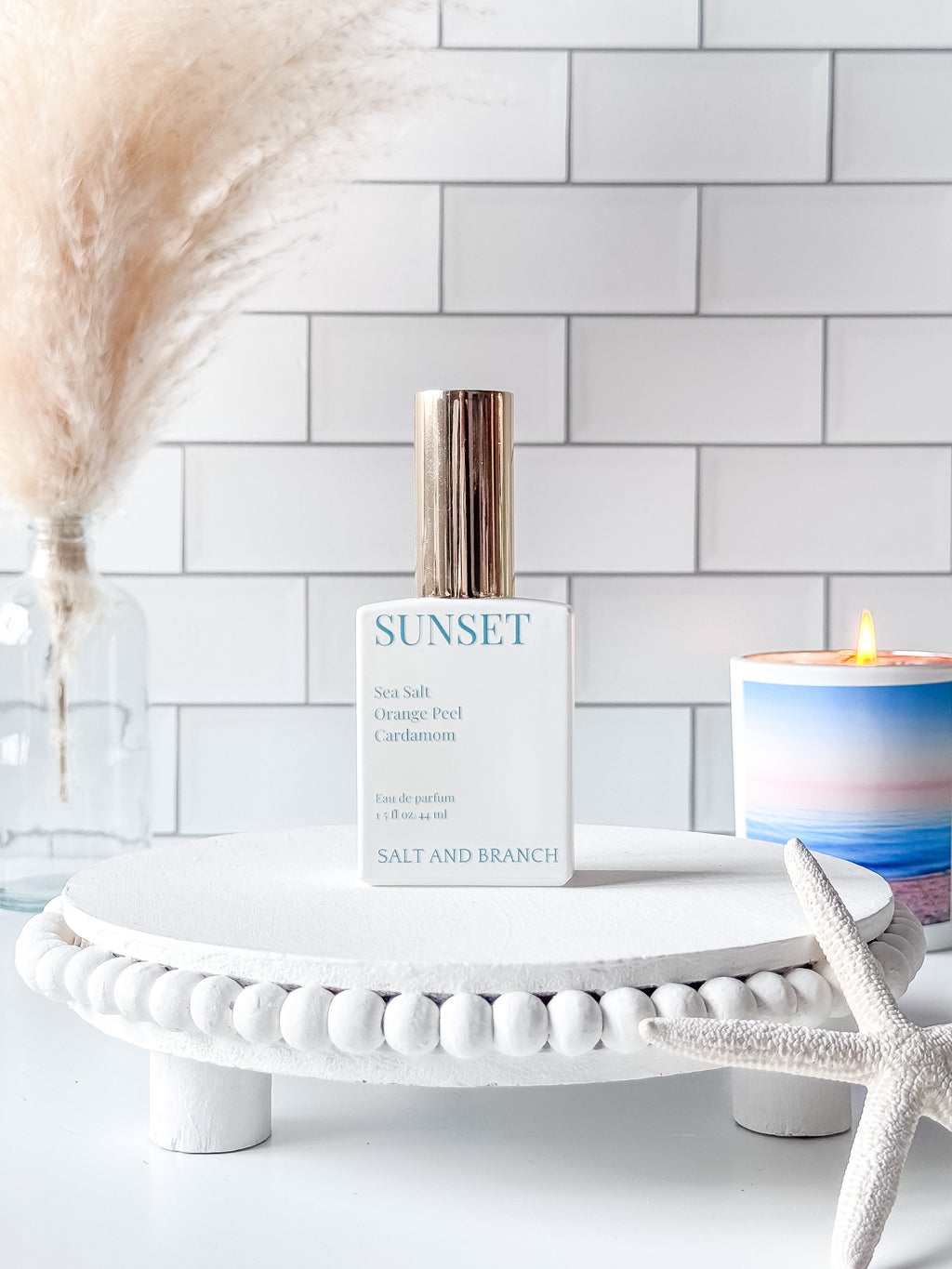 Sunset perfume bottle on a white stand with a candle and decorative elements against a tiled wall.