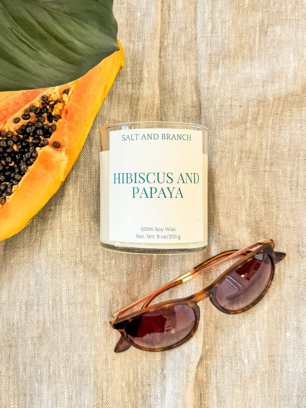 Candle labeled 'Hibiscus and Papaya' by Salt and Branch with a papaya and sunglasses on a textured surface.